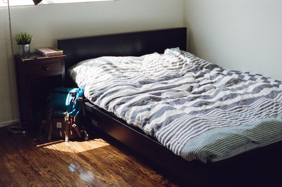 Original photo for test 2: furnished bedroom redesign: Furnished bedroom (bed, left nightstand with plant, book and lamp, backpack, window, dark hardwood with sun patches)